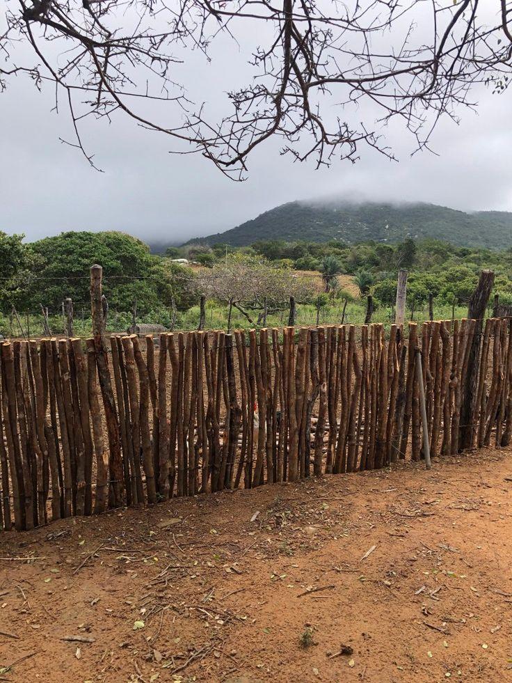 Foto colorida de um terreno de chão de terra batida delimitado por uma cerca de faxina construída com varas de madeira lado a lado. Além da cerca vê-se a paisagem natural, com árvores, montanhas e nuvens