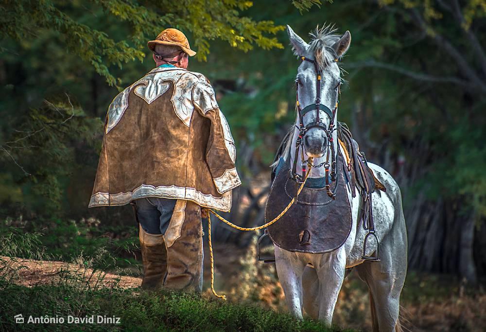 Foto colorida de um homem de costas vestindo uma roupa de couro marrom com detalhes brancos, consistindo em um chapéu em formato de cuia com uma aba curta, uma espécie de jaquetão sobre os ombros que é o gibão, uma proteção de couro que lembra uma calça sem o fundo que ele usa por cima das pernas de uma calça jeans que é a perneira. Ao seu lado de frente para a câmera está um cavalo acinzentado que usa uma proteção de couro escuro no peito. Ao fundo aparece uma mata desfocada.